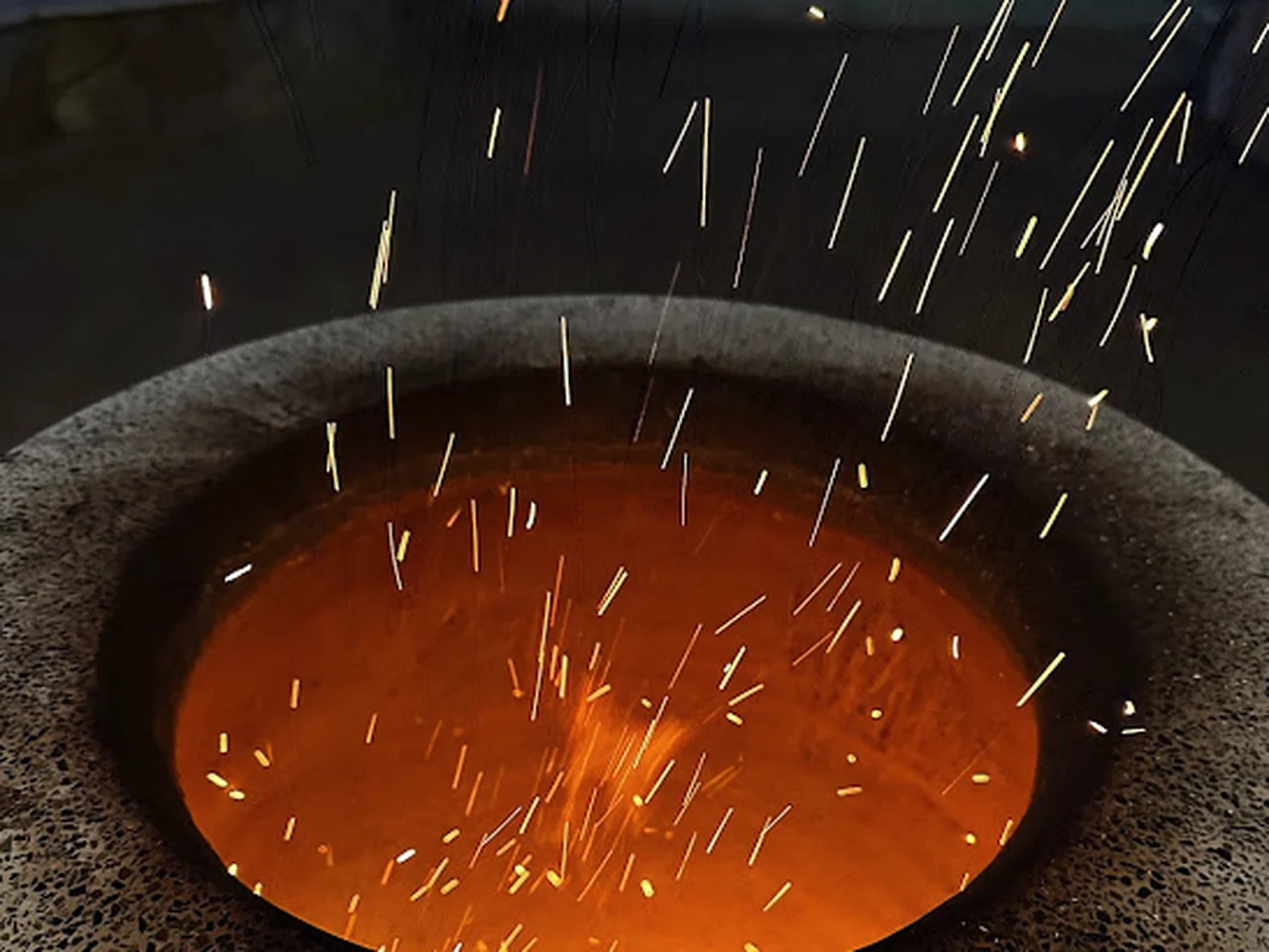 The open mouth of a tandoor filled with glowing orange charcoal, sparks rising into the dark above it.